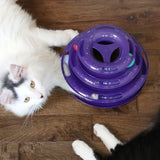 A white cat with black markings lounges on the floor beside a Quirky Kitty Three Layer Ball Track Cat Toy by Quirky Kitty, while a black cats paw is partially visible, suggesting playful interaction.