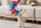 A fluffy kitten stands on its hind legs, playing with the Quirky Kitty Sassy Strawpurry Super Feather Wand. Another cat relaxes on a white sofa in the cozy, bright room with wooden flooring.