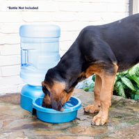 A black and tan dog drinks from a Petmate Big Blue Bowl on a patio. The bowl features a large, clear bottle excellent for outdoor pet hydration. In the background, the wall and greenery are visible, with text reading, Bottle Not Included.