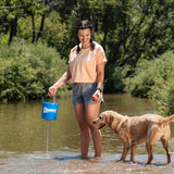A woman in a light peach shirt and gray shorts stands in a creek using her Chuckit! Bucket With Ultra Balls by Chuckit. Beside her, a wet golden retriever waits eagerly to play fetch amid the lush green foliage backdrop.