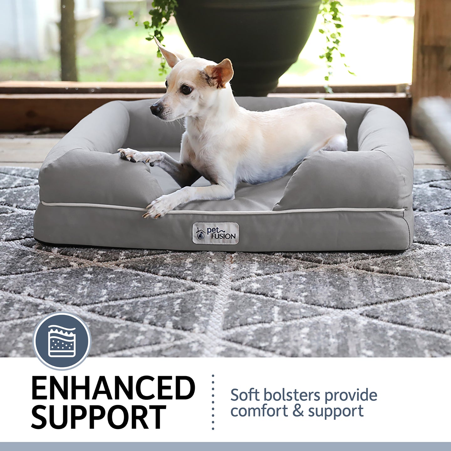 A small dog rests on a PetFusion Ultimate Dog Bed with gray memory foam and raised bolsters for joint pain relief. Set outdoors on a porch with plants, a label states: Enhanced Support: Soft bolsters provide comfort & support.