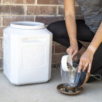 Kneeling on the ground in black leggings and a gray top, a person scoops birdseed into a small plastic container on a saucer. Nearby, against the brick wall, stands the Vittles Vault Bird Seed Storage Container by Vittles Vault with floral designs for secure seed storage.