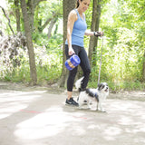 A woman in athletic wear walks her small black and white dog through a forest, holding a blue leash and a Vittles Vault Travel-Trainer Portable Food Storage Container. Sunlight filters through the green trees.