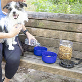 A person cradles a small black and white dog, pouring water from a BPA-free bottle into a blue bowl on a wooden bench. Next to an airtight Vittles Vault Travel-Trainer Portable Food Storage Container is another blue bowl filled with kibble, all set in a lush green outdoor setting.