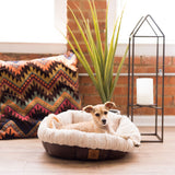 A small tan dog lies comfortably in a SnooZZy Rustic Luxury Ultra Cuddler Pet Bed, featuring a plush, round design and non-skid bottom. The setting includes a colorful cushion, tall green plant, and candle in a metal holder against a softly lit brick wall.