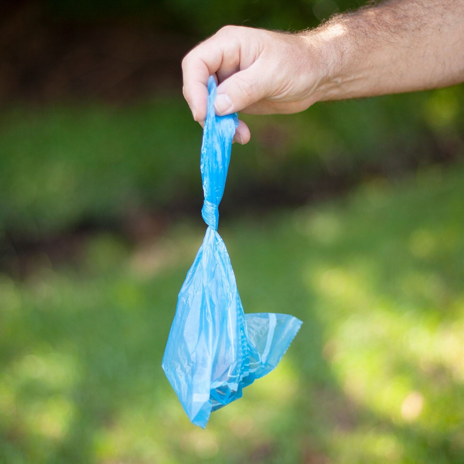 A hand holds an Arm & Hammer Easy-Tie Dog Waste Bag, designed for odor control, against a blurred green outdoor background.