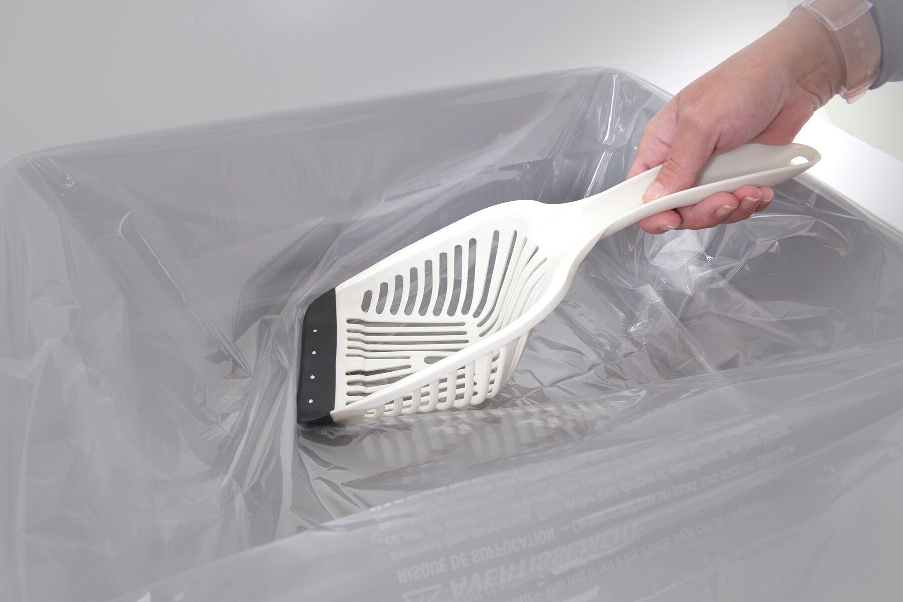 A hand sifts through a gray kitty litter box lined with a transparent plastic bag using the Aspen Pet No Tear Cat Litter Scoop, featuring a comfortable grip.