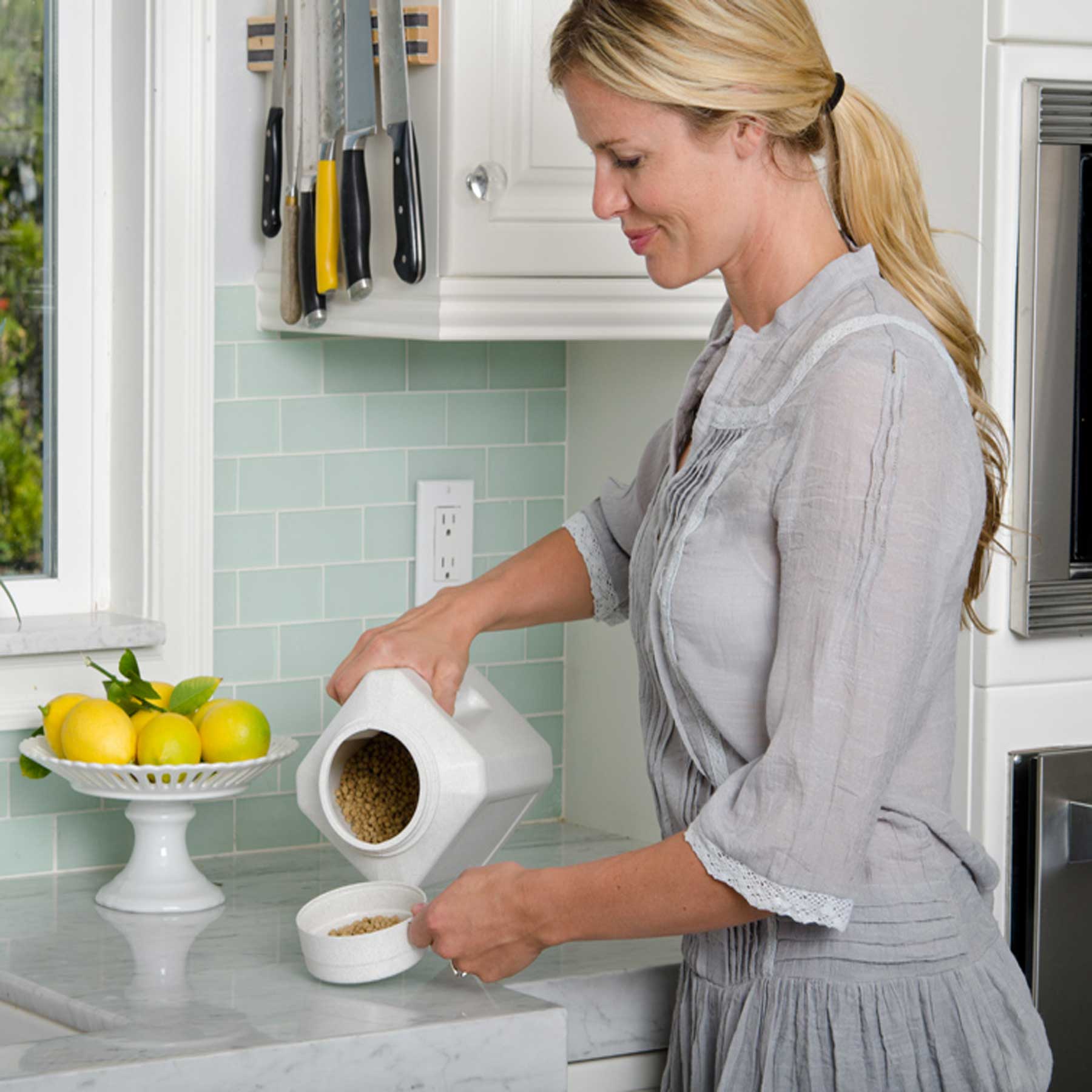 A woman in a light gray blouse pours grains from a white container, similar to the BPA-free Vittles Vault Junior by Vittles Vault, into a small white bowl on the kitchen counter. To her left is a bowl of lemons, and knives hang on a magnetic strip in the background.